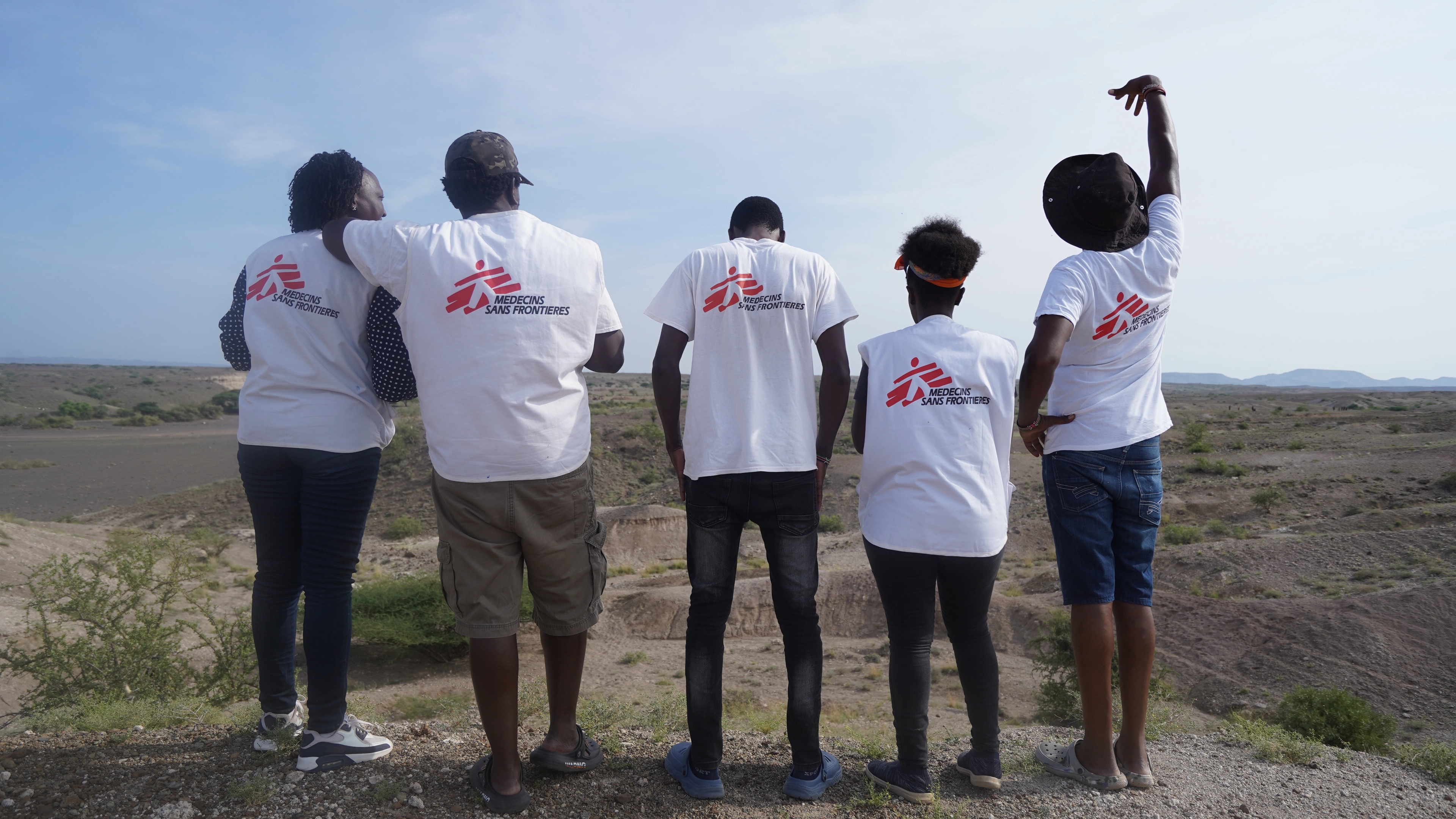 Five people in MSF shirts and vests stand with their backs to the camera, one has an arm raised