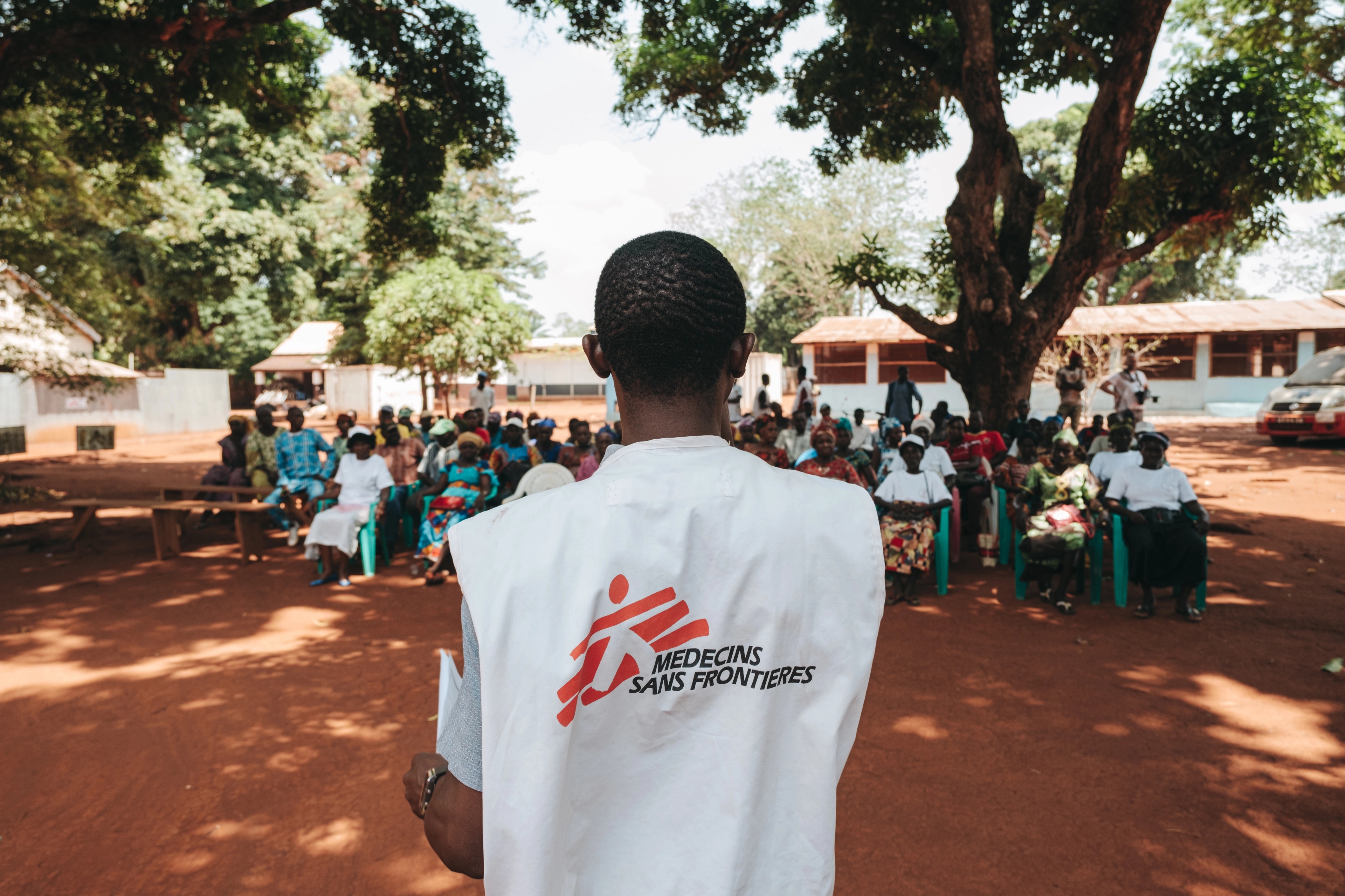 A man in a white MSF branded vest has his back towards the camera, and speaks to a small audience of people seated outdoors