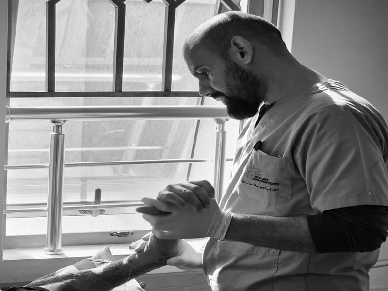 A man stands in scrubs and holds a patient's hand, in black and white